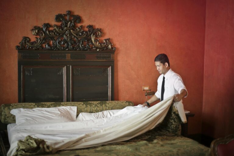 Side view of young ethnic male making bed while cleaning stylish hotel room with vintage furniture and red walls